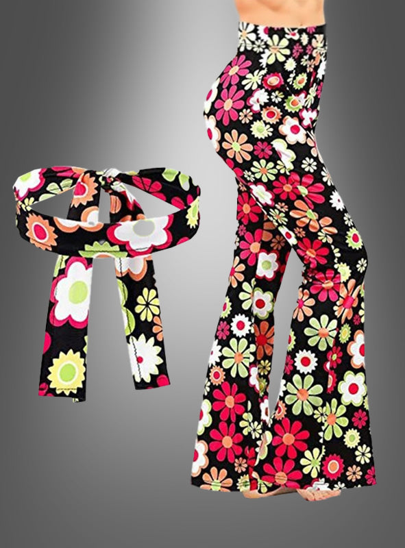 Frau in der Schlaghose „Hippie Set Flowerful“ – eine hoch taillierte Schlaghose mit passendem breitem Gürtel, verziert mit leuchtenden Blumen auf schwarzem Hintergrund – perfekt als 70er-Jahre-Damen-Schlaghose für Ihr nächstes Hippie-Party-Outfit.