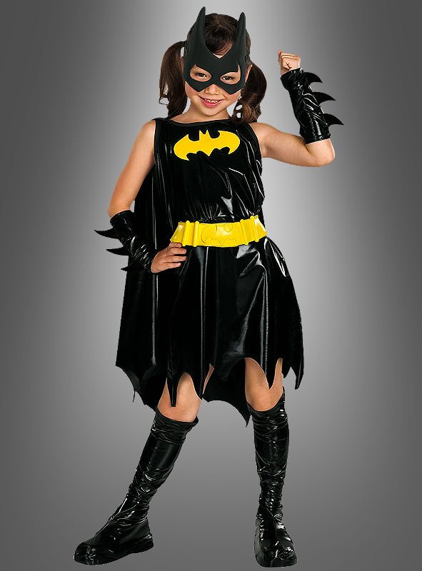 Ein Kind in einem „Batgirl Deluxe“-Kinderkostüm in Schwarz und Gelb posiert mit erhobenem Arm wie eine echte Superheldin. Vor einem grauen Hintergrund trägt sie eine Maske, einen Umhang, einen Gürtel, Armbänder und Stiefel.