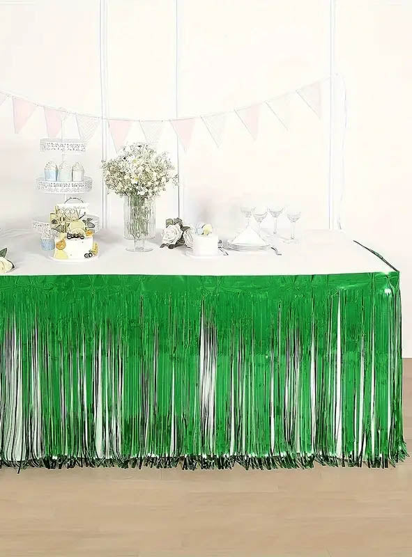 Ein für eine Party geschmückter Tisch wird durch den glänzend grünen „Lametta Tischrock“ (75 x 275 cm) als Tischumrandung geprägt, der mit Blumen, Glaswaren und einem Etagerenständer verziert ist. Eine weiße Wand und pastellfarbene Wimpelketten im Hintergrund runden die bezaubernde Partydekoration ab.