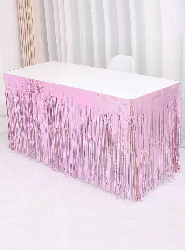 Ein rechteckiger Tisch mit weißer Tischplatte ist an den Seiten mit dem „Lametta Tischrock 75 x 275 cm“ in glänzendem Rosa verziert und steht vor weißen Vorhängen auf einem weißen Teppich, wodurch eine festliche Partydekoration entsteht.