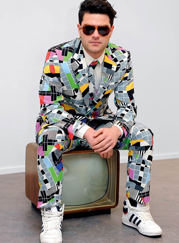 Ein Mann in einem farbenfrohen „Deluxe Testbild“-Anzug von OppoSuits mit geometrischem Muster und Sonnenbrille sitzt auf einem alten Fernseher und trägt weiße Sneaker vor einem schlichten Hintergrund – ideal für eine 70er-Jahre-Mottoparty oder als auffälliges Karnevalskostüm.
