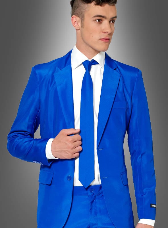Ein Mann, gekleidet in einen blauen Anzug von „Anzugmeister“, ergänzt durch ein strahlend weißes Hemd und eine blaue Krawatte, posiert vor einem grauen Hintergrund mit Farbverlauf.