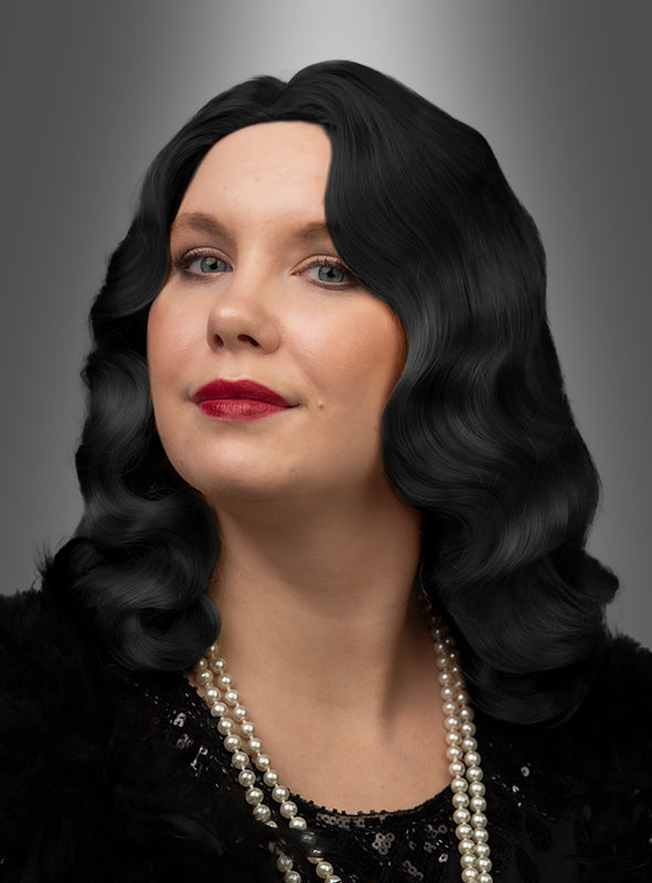 Eine Frau mit welligem schwarzem Haar, das mit der Perücke „20er Jahre Premium Wasserwelle Charlotte“ gestylt ist, posiert in einem schwarzen Oberteil, mit mehreren Perlenketten und rotem Lippenstift vor einem grauen Hintergrund mit Farbverlauf.