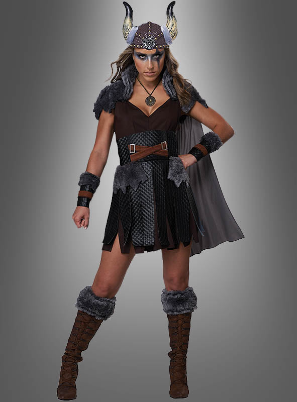 Eine Frau steht mit dem Gesicht nach vorne vor einem grauen Hintergrund mit Farbverlauf. Sie trägt das von der Wikingertradition inspirierte „Sexy Wikingerin Schildmaid“-Kostüm, bestehend aus einem gehörnten Helm, einem braunen Kleid mit Pelzdetails, passenden Armbändern, Stiefeln und einem Umhang.