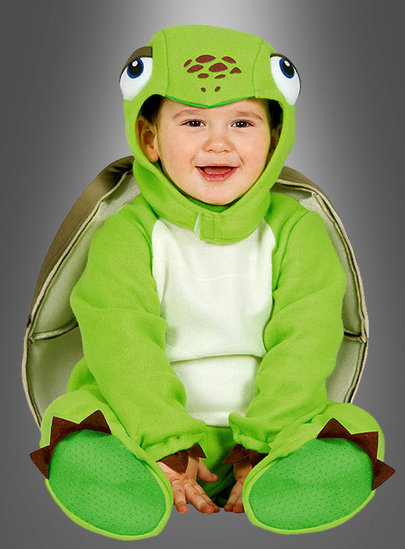 Auf dem Bild ist ein fröhliches Kind zu sehen, das vor einem grauen Hintergrund sitzt und das „Baby-Schildkröte-Kostüm“ trägt – ein entzückendes grünes Schildkrötenkostüm mit einer Kapuze mit großen Comic-Augen, einem Panzer auf dem Rücken und flossenförmigen Füßen.