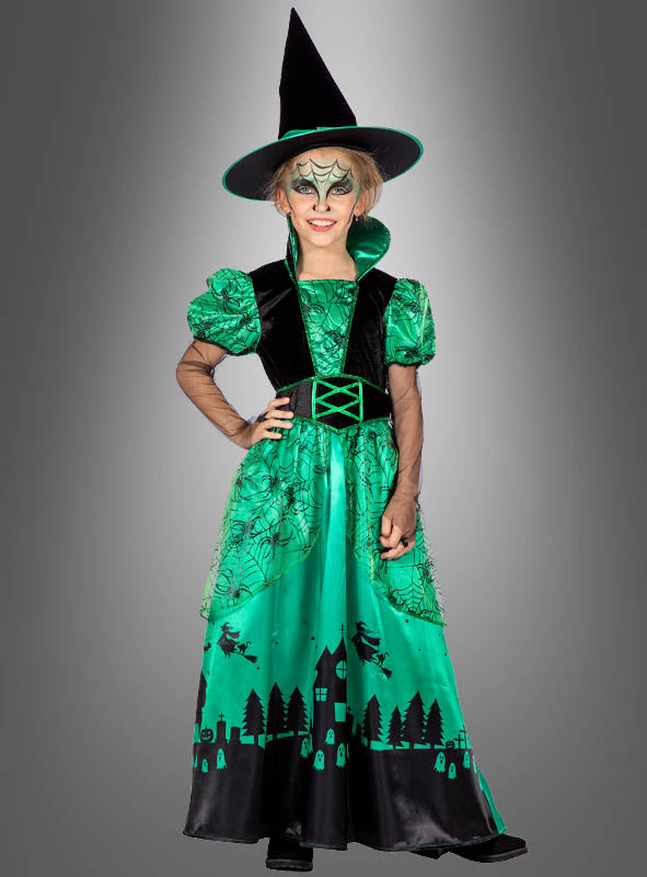 Ein Kind, das das „Langes Hexenkleid grün für Kinder“ trägt – ein grün-schwarzes Hexenkostüm mit spitzem Hut, Spinnennetz-Make-up und einem Kleid, das mit Halloween-Motiven verziert ist –, steht vor einem grauen Hintergrund mit Farbverlauf. Dieses bezaubernde Outfit fängt die Magie des Halloween-Hexenkostüms für Kinder perfekt ein.