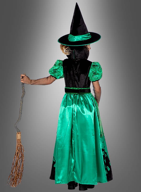 Ein Kind, das das Kostüm „Langes Hexenkleid grün für Kinder“ mit einer spitzen Mütze trägt und einen Besenstiel in der Hand hält, steht mit dem Rücken zur Kamera vor einem grauen Hintergrund.
