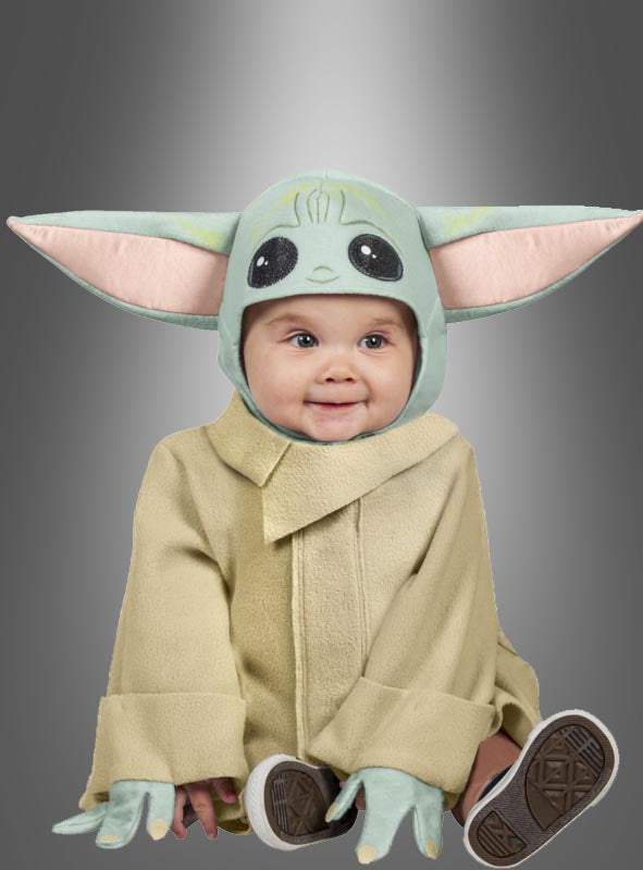 Ein Baby, das das „Baby-Yoda-Kostüm ‚Das Kind‘ Grogu aus Star Wars“ trägt – mit großen grünen Ohren und einem hellbraunen Gewand –, sitzt vor einem grauen Hintergrund mit Farbverlauf und lächelt leicht.
