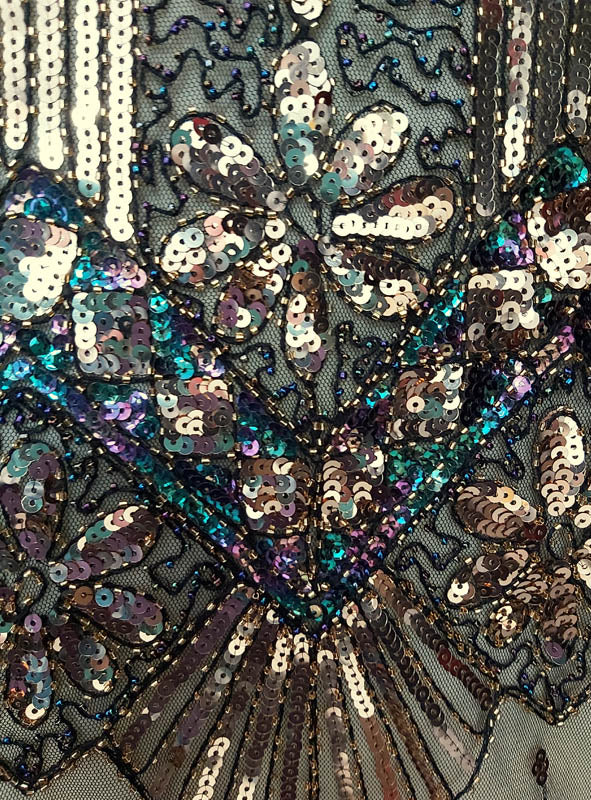 Nahaufnahme eines Stoffes, der mit schillernden Paillettenmustern in floralen und geometrischen Motiven auf einem Netzhintergrund bestickt ist und an die „Luxus 1920er Jahre Kleider Shirley“ erinnert.