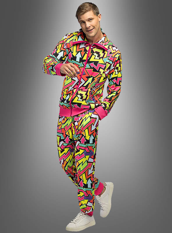Ein Mann, der den „Cooler Trainingsanzug Neon 80er Flash Herren“ trägt – einen farbenfrohen Trainingsanzug mit bunten, geometrischen Formen in Pink, Gelb und Grün, die an ein kultiges 80er-Jahre-Kostüm erinnern –, steht vor einem grauen Hintergrund mit Farbverlauf und lächelt.