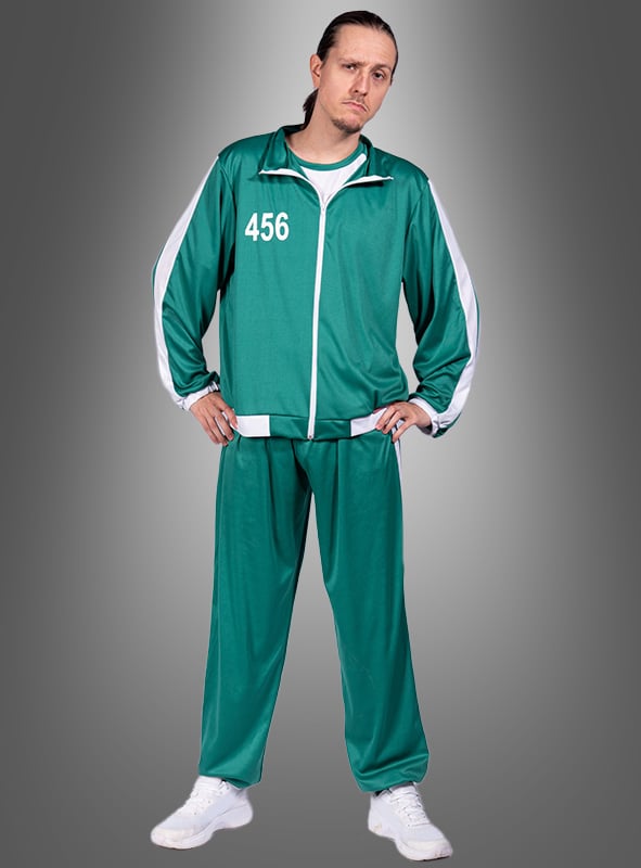 Eine Person trägt den „Game Spieler 456“ Trainingsanzug für Herren in Grün mit der Nummer 456, dazu weiße Turnschuhe, und steht vor einem grauen Hintergrund mit Farbverlauf.