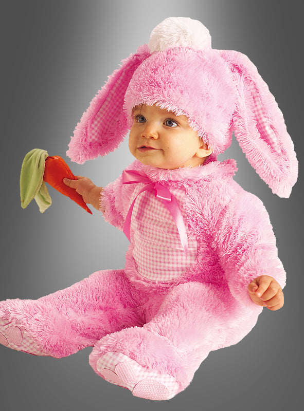 Ein Baby, das den „Baby-Hase ROSA Strampelanzug“ trägt – ein rosa Hasenkostüm mit Schlappohren und einer weißen Bommelmütze –, hält eine Plüschkarotte in der Hand, während es vor einem grauen Hintergrund mit Farbverlauf sitzt.