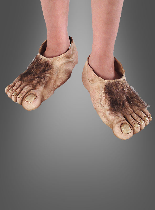 Ein Kind trägt große, realistisch gestaltete Hobbit-Schuhüberzieher für Kinder mit übertrieben geformten Zehen und Kunsthaar, die den übergroßen Hobbit-Füßen aus „Der Herr der Ringe“ ähneln, vor einem schlichten grauen Hintergrund.