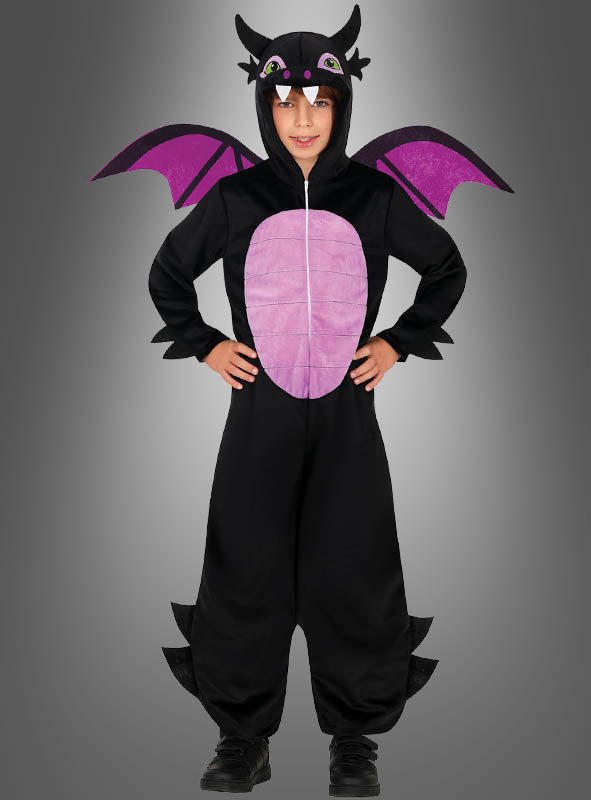 Ein Kind steht mit den Händen in den Hüften und trägt einen „Schwarzer Drachen“-Overall – ein schwarz-lila Drachenkostüm mit Flügeln, Hörnern und einem hellrosa Bauch – vor einem grauen Hintergrund mit Farbverlauf.