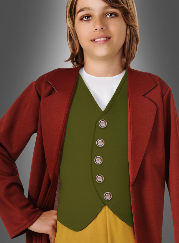Ein Kind mit schulterlangen Haaren steht vor einem grauen Hintergrund und trägt das Bilbo-Hobbit-Kinderkostüm – ein bezauberndes Ensemble, das von „Der Herr der Ringe“ inspiriert ist und aus einem roten Mantel, einer grünen Weste mit Knöpfen und einer gelben Hose besteht, die Bilbos ikonischen Hobbit-Look perfekt verkörpern.