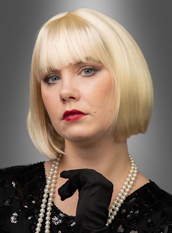 Eine Frau präsentiert die hochwertige Pagenschnitt-Bob-Perücke „Vera“ mit kurzem blondem Haar im klassischen Bob-Stil. Sie trägt ein schwarzes Oberteil und schwarze Handschuhe, dazu eine Perlenkette und roten Lippenstift, und posiert vor einem grauen Hintergrund mit Farbverlauf.
