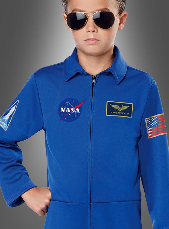 Ein Kind, das den blauen Astronautenanzug der NASA für Kinder trägt – mit Missionsabzeichen, Pilotenbrille und einem Aufnäher mit der amerikanischen Flagge –, steht vor einem grauen Hintergrund mit Farbverlauf.