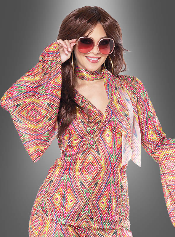 Eine Person trägt einen bunten Disco-Anzug mit Pailletten für Damen mit Schlaghose und einer großen Sonnenbrille und posiert, wobei sie die Brille mit einer Hand vor einem grauen Hintergrund mit Farbverlauf hält.