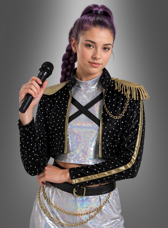 Eine Frau mit violetten Zöpfen hält ein Mikrofon in der Hand und trägt das K-Pop-Star-Kostüm „Romy Sängerin“ – ein funkelndes silbernes Kostüm, kombiniert mit einer schwarzen Nietenjacke mit goldenen Verzierungen und Kettenelementen –, während sie vor einem grauen Hintergrund mit Farbverlauf steht.
