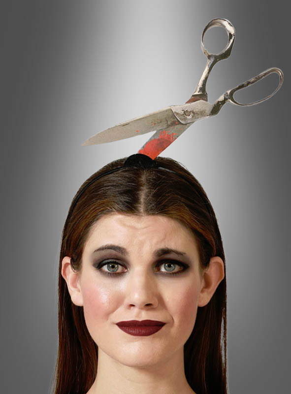 Eine Frau mit dunklem Make-up trägt das Halloween-Accessoire „Horror Haarreif Werkzeuge im Kopf“, ein Stirnband, das so gestaltet ist, dass es aussieht, als stecke eine große Schere durch ihren Kopf; im Hintergrund ist ein grauer Farbverlauf zu sehen.