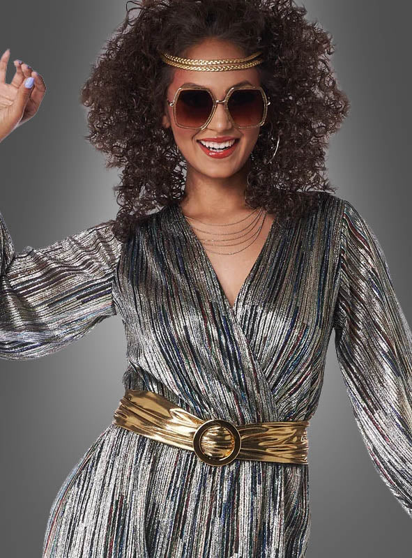 Eine Frau, die den „Disco-Kostüm-Jumpsuit Super Nova“ trägt – einen glänzenden Jumpsuit in Metallic-Optik –, kombiniert mit einer großen Sonnenbrille, einem goldenen Gürtel und einem goldenen Stirnband, posiert lächelnd vor einem grauen Hintergrund.