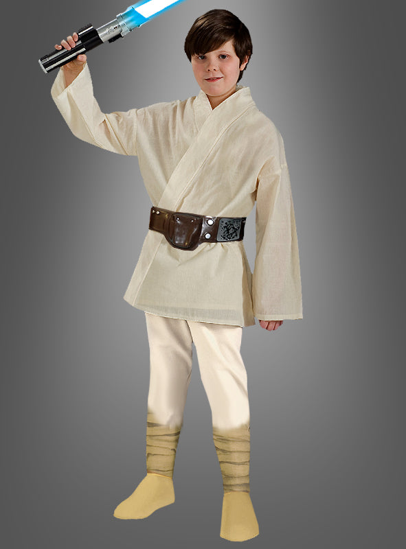 Ein Kind in einem „Deluxe Luke Skywalker“-Star-Wars-Kinderkostüm hält ein blaues Lichtschwert in der Hand und steht vor einem schlichten grauen Hintergrund.