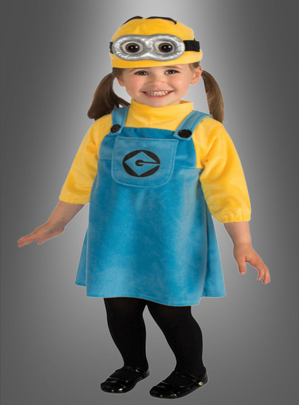 Ein kleines Kind, das das Original-Minion-Mädchen-Kostüm trägt – bestehend aus einem blauen Strickkleid, einem gelben Shirt, schwarzen Strumpfhosen, schwarzen Schuhen und einer Minion-Augenmaske –, steht lächelnd vor einem grauen Hintergrund.