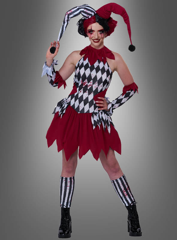Eine Person im Kostüm „Verrückte Harlekin-Dame Topsy“ – lächelnd in einem rot-schwarzen Narrenkostüm mit Gesichtsbemalung, gestreiften Handschuhen und Socken sowie einer klassischen Narrenmütze – vor einem grauen Hintergrund.