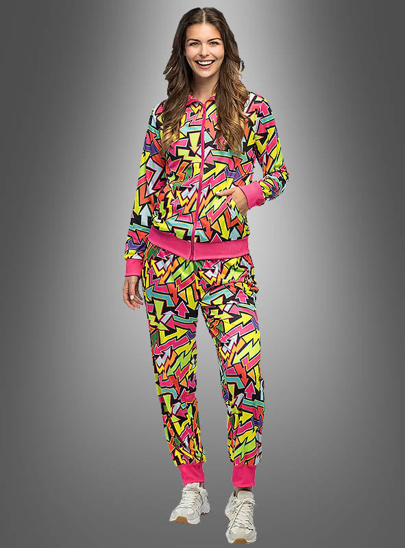 Eine Person steht lächelnd vor einem grauen Hintergrund mit Farbverlauf und trägt den UV-Trainingsanzug „Neon Flash“ für Damen – einen farbenfrohen Trainingsanzug mit geometrischen Mustern, rosa Akzenten und weißen Sneakers. Dieser lebhafte Look fängt den Geist eines Retro-Kostüms aus den 80ern perfekt ein und eignet sich ideal als Damen-Retro-Kostüm für jedes 80er- und 90er-Party-Outfit.