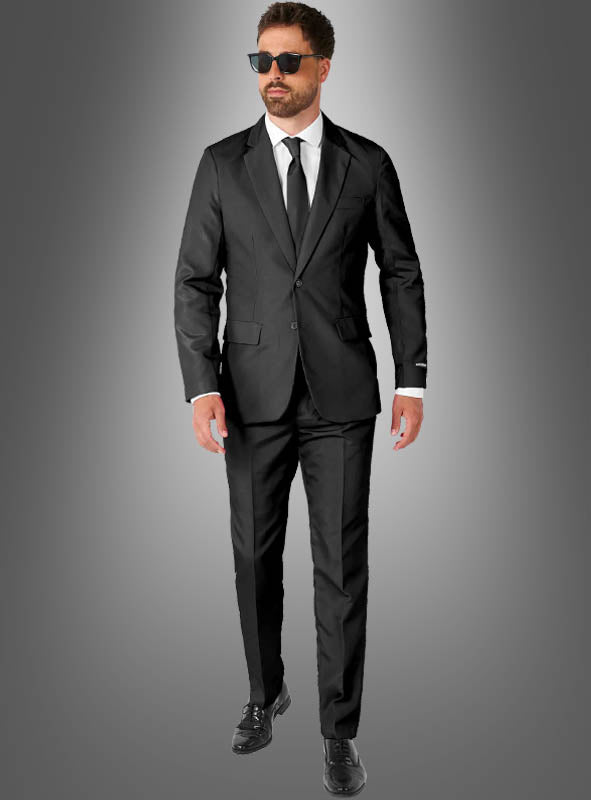 Ein Mann trägt einen schwarzen Anzug mit Krawatte von Suitmeister, kombiniert mit einem weißen Hemd, schwarzen Schuhen und einer Sonnenbrille, und steht vor einem grauen Hintergrund mit Farbverlauf.