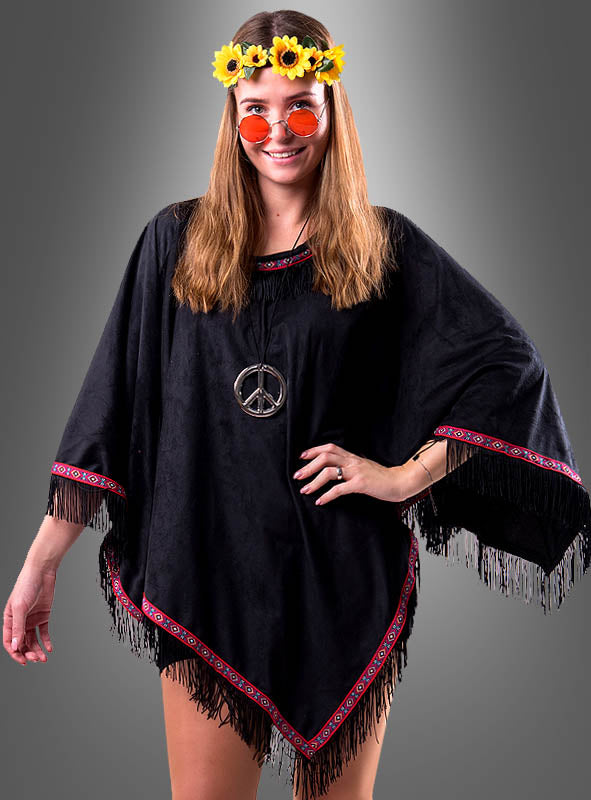 Eine Frau trägt den „Wandelbaren Poncho mit Fransen Hippie Indianer“ – aus schwarzem Stoff mit roter Borte –, kombiniert mit einer Halskette mit Peace-Zeichen, einer runden orangefarbenen Sonnenbrille und einem gelben Blumenstirnband. Dieser vielseitige Look eignet sich perfekt für Damen und Herren und kommt vor einem grauen Hintergrund besonders gut zur Geltung.