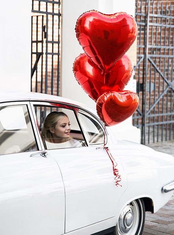 Eine Frau sitzt auf dem Rücksitz eines weißen Oldtimers und hält drei „Herz-Folienballons – Geschenk für viele Anlässe“ aus dem Fenster in der Nähe eines Metalltors.