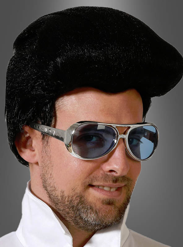 Ein Mann mit einer schwarzen Pompadour-Perücke, gekleidet in ein weißes Outfit mit Kragen, posiert vor einem grauen Hintergrund und trägt dabei die Pilotenbrille „King of Rock Elvis Brille 60er“.