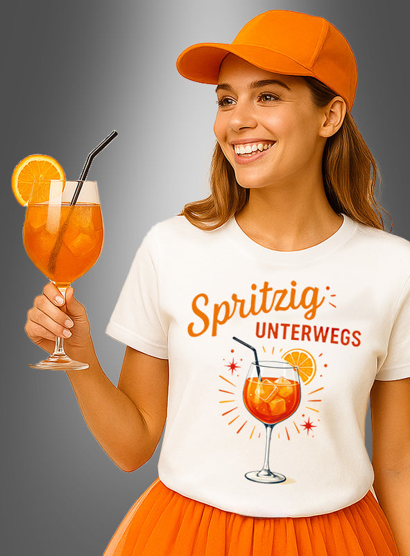 Die Frau lächelt und hält einen mit einem Strohhalm und einer Orangenscheibe garnierten Cocktail in der Hand. Sie trägt das Cocktail-T-Shirt „Spritzig unterwegs“ – mit dem verspielten Slogan „Spritzig unterwegs“ und einer passenden Getränkeillustration. Dieses leuchtend orangefarbene Shirt eignet sich ideal als Sommer-Gruppenshirt für festliche Anlässe.