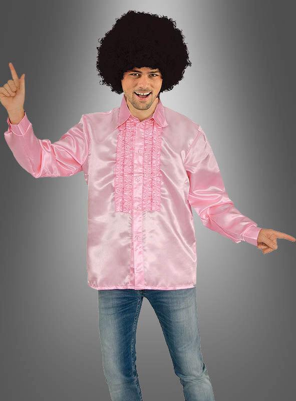 Ein Mann mit einer lockigen schwarzen Perücke, einem glänzenden rosa Herrenhemd im 70er-Jahre-Stil und blauen Jeans zeigt vor einem grauen Farbverlaufshintergrund nach oben und zur Seite – und verkörpert damit den ultimativen Disco-Look für Herren.