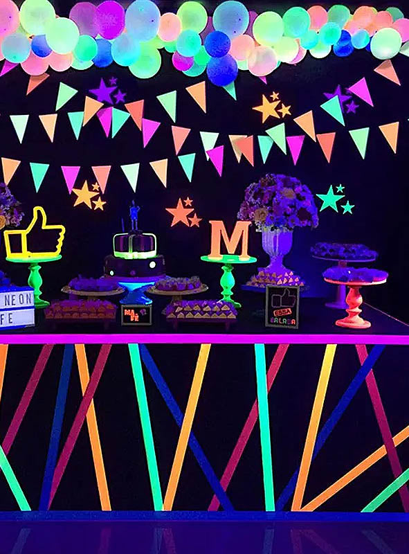 Ein Party-Tisch im Neon-Stil mit leuchtenden Dekorationen, Kuchen, Luftballons und Girlanden wird vollständig von Schwarzlicht beleuchtet. Leuchtende Formen und Schilder unterstreichen die lebhafte, festliche Gestaltung, während die „80er Jahre Neon-Girlanden 440 cm Deko“ für einen zusätzlichen Farbtupfer sorgt – perfekt für jede Schwarzlicht-Party-Dekoration.