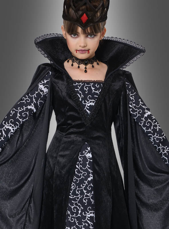 Ein Kind, das das „Schwarze Vampir-Königin“-Kinderkostüm „Lina“ trägt – ein dramatisches schwarz-weißes Vampir-Königin-Kostüm für Kinder mit auffälligem Kragen, Krone und einem schwarzen Vampir-Kleid –, steht vor einem grauen Hintergrund; für einen zusätzlichen Effekt wurde ihm Kunstblut um den Mund herum aufgetragen.