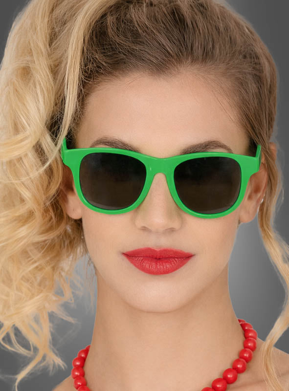Eine Frau mit welligem blondem Haar trägt eine leuchtend grüne Sonnenbrille im Wayfarer-Stil in verschiedenen Farben, roten Lippenstift und eine rote Perlenkette vor einem grauen Hintergrund.