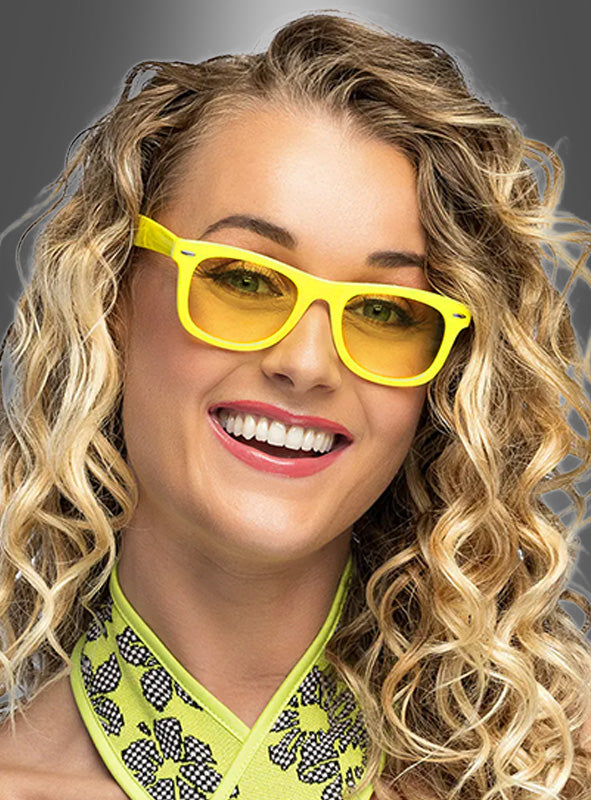 Eine Frau mit lockigem blondem Haar lächelt vor einem grauen Hintergrund mit Farbverlauf in die Kamera; sie trägt eine gelbe Sonnenbrille im Wayfarer-Stil von „Party Brille“ in verschiedenen Farben sowie einen grün gemusterten Schal.