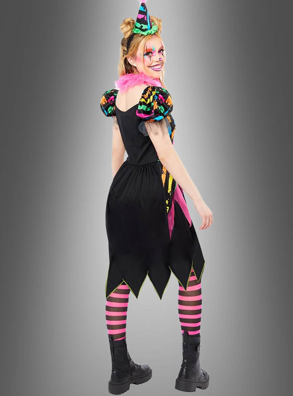 Eine Person, die ein farbenfrohes „Zirkus Clown Neon“-Kostüm für Damen trägt, dazu gestreifte rosa-schwarze Strumpfhosen, schwarze Stiefel und einen Partyhut, steht vor einem grauen Hintergrund und lächelt.