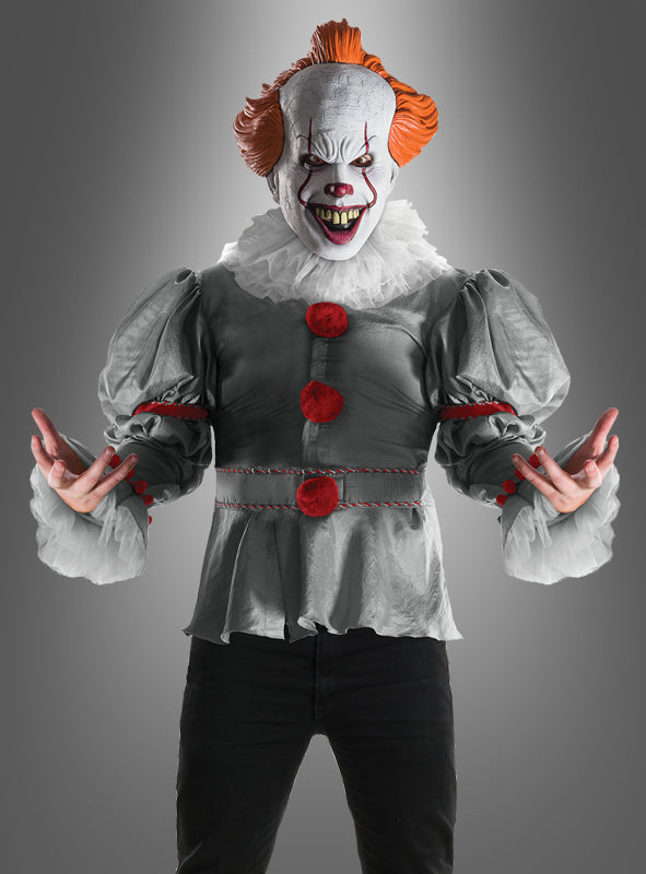 Eine Person, die das „Horrorfilm ES“-Kostüm von Pennywise trägt – mit weißem Make-up, einer roten Nase, orangefarbenen Haaren, Puffärmeln und roten Akzenten –, steht mit ausgestreckten Armen vor einem grauen Hintergrund.