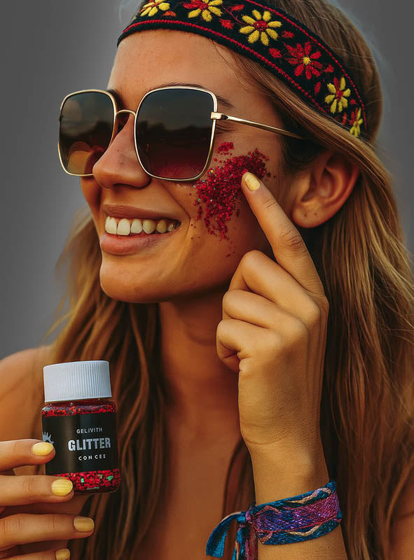 Eine Frau mit Sonnenbrille und einem geblümten Stirnband trägt mit dem Finger rotes Glittergel-Make-up in verschiedenen Farben auf ihre Wange auf. Sie lächelt und hält einen kleinen Behälter mit der Aufschrift „Glitter“ in der Hand.