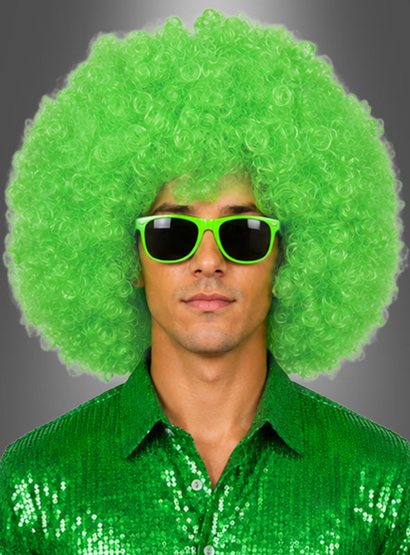 Ein Mann trägt eine XXL-Riesen-Afro-Perücke in leuchtendem, lockigem Grün, dazu eine grüne Sonnenbrille und ein glänzendes grünes Paillettenhemd, vor einem neutralen Hintergrund.