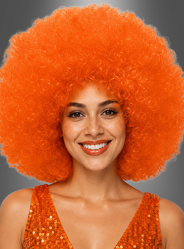 Eine lächelnde Person, die eine orangefarbene XXL-Riesen-Afro-Perücke in tollen Farben und ein orangefarbenes Pailletten-Top trägt und vor einem grauen Hintergrund posiert.