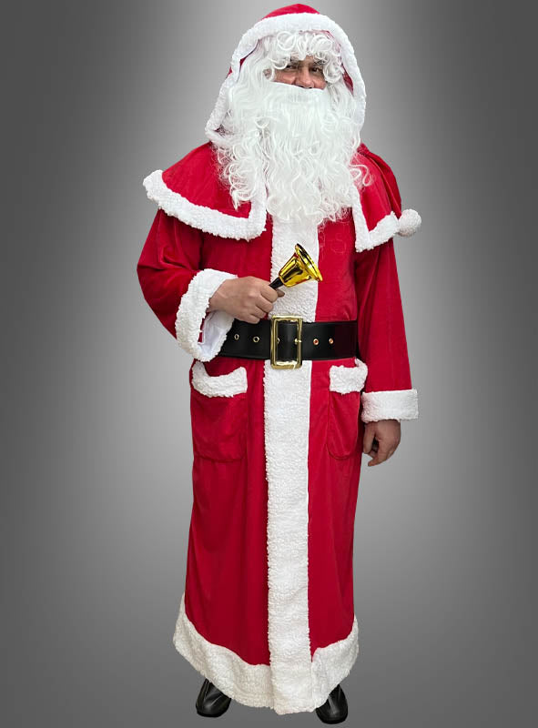 Eine Person, die einen Weihnachtsmann-Mantel mit Stiefelüberzügen trägt, einen weißen Bart und ein rotes Plüschkostüm hat und eine kleine goldene Glocke in der Hand hält, während sie vor einem grauen Hintergrund steht.