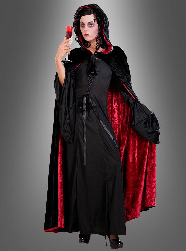Vor einem grauen Hintergrund steht eine Person, die das „Premium Kapuzen Wendecape Unisex“ in auffälligem Schwarz und Rot trägt, dazu ein langes schwarzes Kleid, und in der Hand hält sie ein Glas, das mit roter Flüssigkeit gefüllt ist.