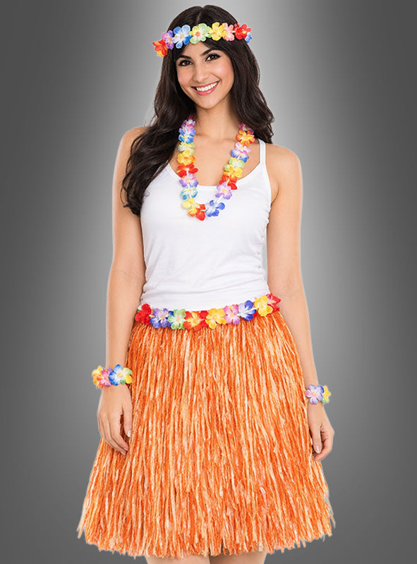Eine Frau steht lächelnd vor einem grauen Hintergrund und trägt ein weißes Trägertop sowie das 5-teilige Hawaii-Set mit Rock und Accessoires, zu dem ein orangefarbener Grasrock und bunte Blumen-Accessoires gehören.