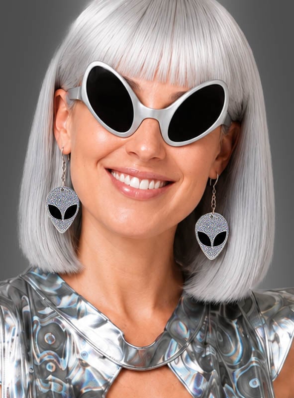 Eine Frau mit silbernem Haar, die eine große schwarze Sonnenbrille und ein metallisches Oberteil trägt, lächelt vor einem schlichten grauen Hintergrund, während sie die „Alien-Ohrringe für Space-Kostüme“ präsentiert – Ohrringe in Form von silbernen Aliens.