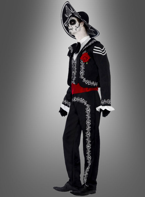 Eine Person, die das „Tag der Toten“-Bräutigam-Kostüm trägt – bestehend aus einer von Mariachi inspirierten Skelett-Gesichtsbemalung, einem schwarzen Hut und einem Anzug, der mit weißer Stickerei, einer roten Schärpe und einer Rose verziert ist –, steht vor einem grauen Hintergrund mit Farbverlauf.