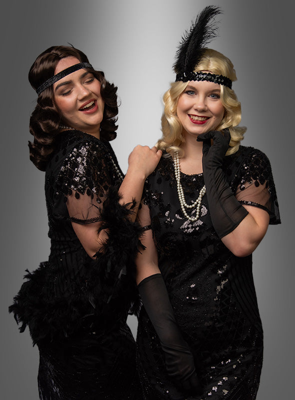 Zwei Frauen in schwarzen Kleidern im Stil der 1920er Jahre, mit Handschuhen und gefiederten Stirnbändern posieren gemeinsam und lächeln vor einem grauen Hintergrund mit Farbverlauf. Ihr Glamour wird durch die eleganten „Luxus 20er Premium Wasserwelle Perücke Elli“-Perücken unterstrichen, die den ikonischen Looks dieser Epoche perfekt entsprechen.